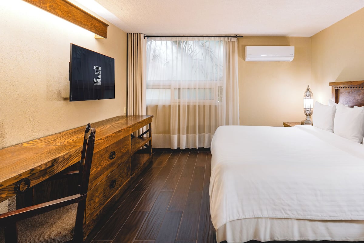 Une chambre d’hôtel avec un lit en bois, un bureau en bois, un téléviseur fixé au mur et une fenêtre avec des rideaux.