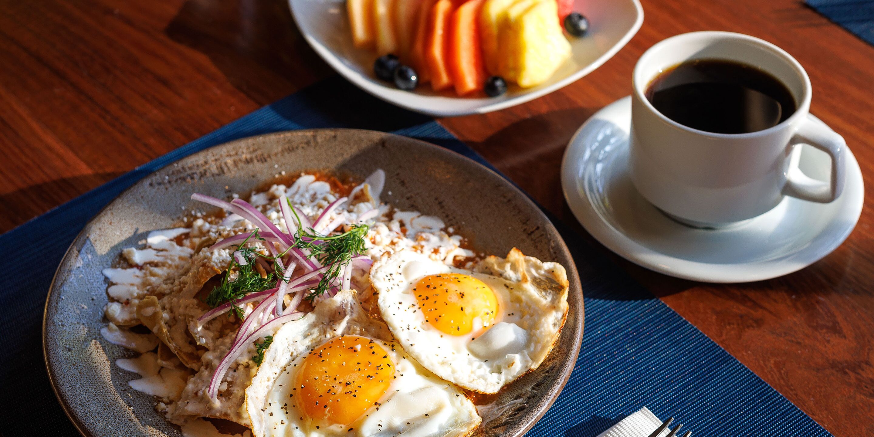 A breakfast plate with eggs, onions, and herbs, a bowl of fruit, and a cup of coffee on a wooden table.