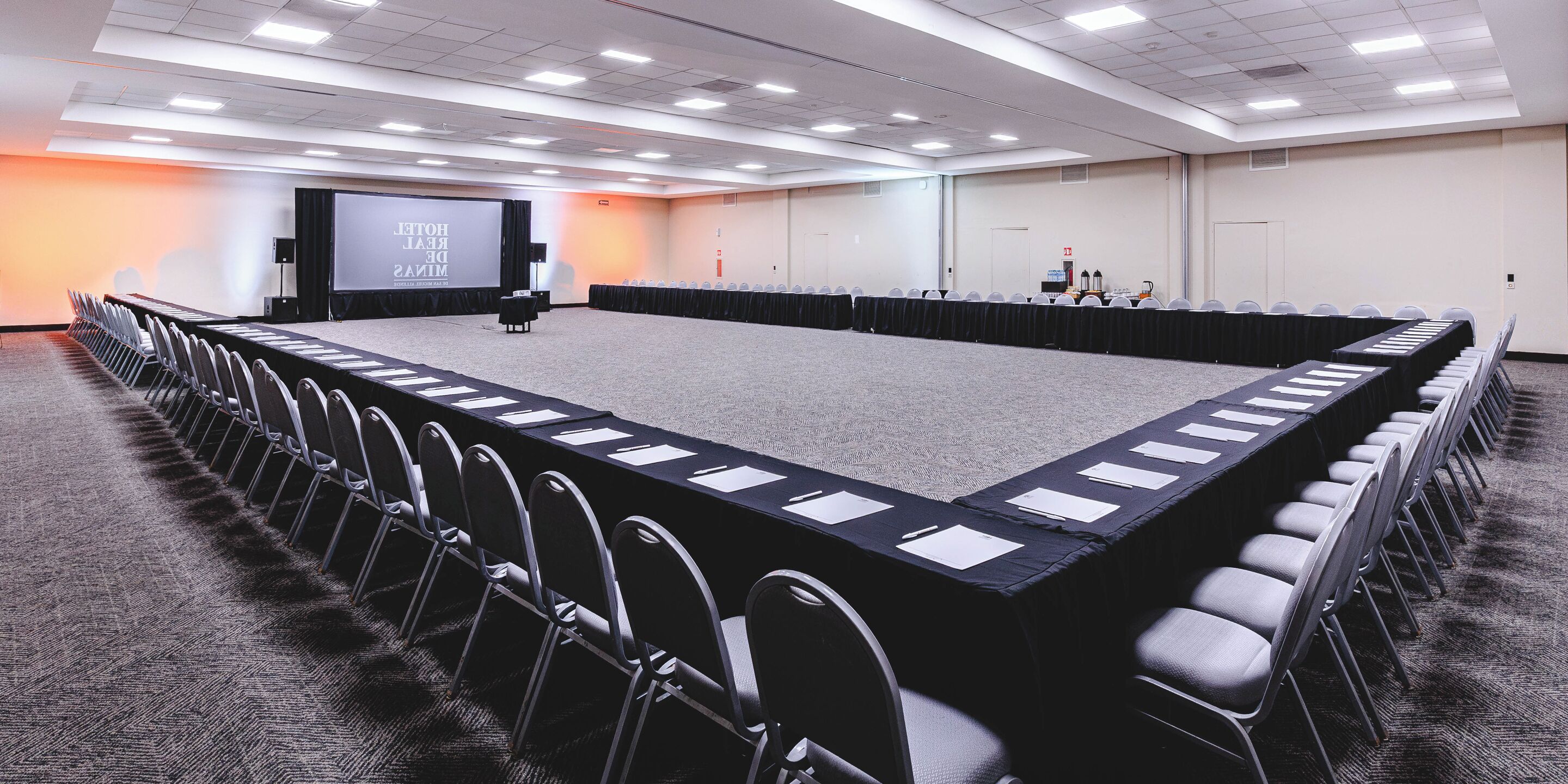 Un salón de conferencias con una larga mesa, sillas, una pantalla de proyector y piso alfombrado.