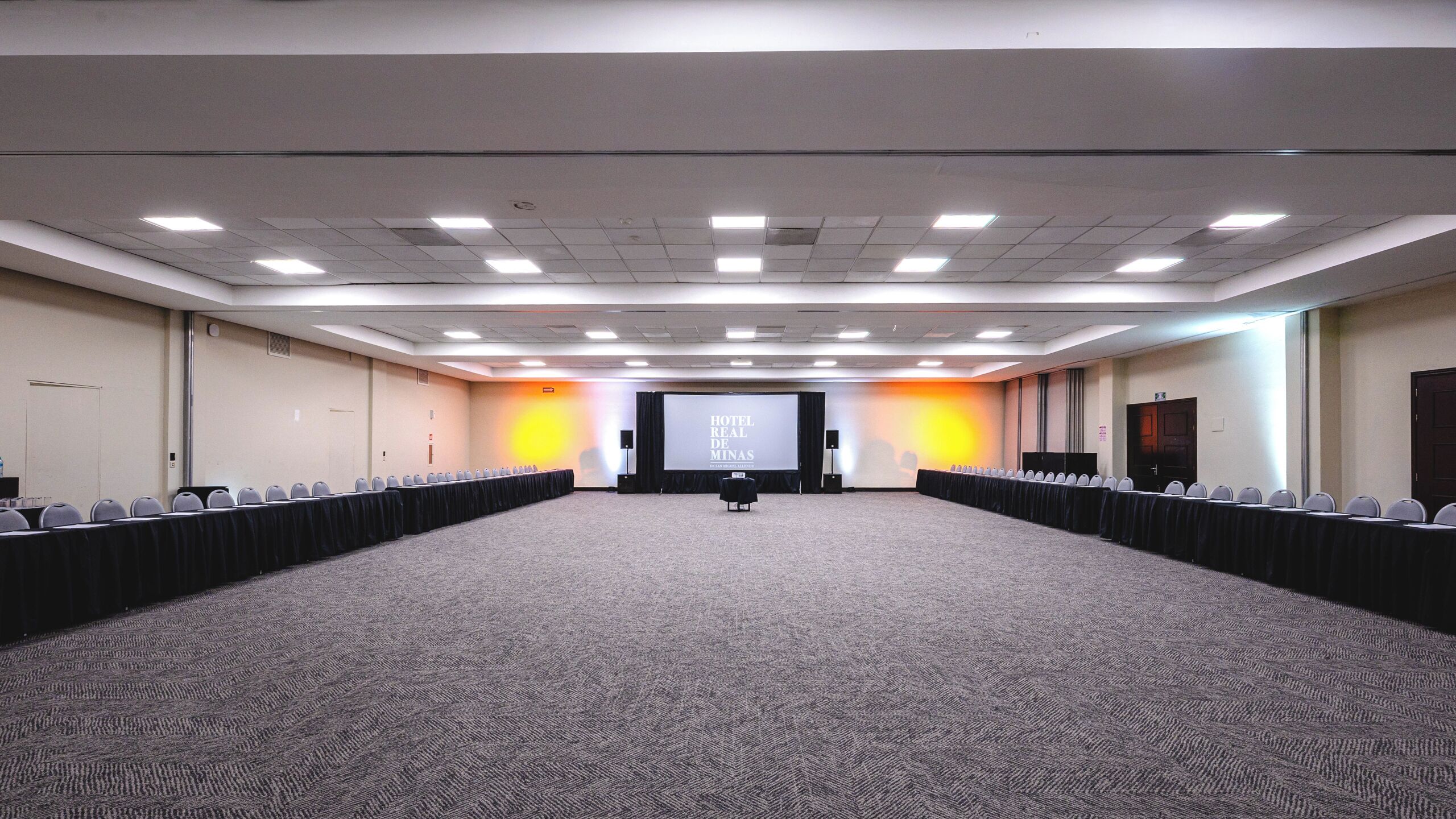 Un amplio salón de conferencias con piso alfombrado, filas de sillas, una pantalla de proyector y una zona para escenario.