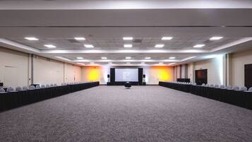 A large conference room with a carpeted floor, rows of chairs, a projector screen, and a stage area.