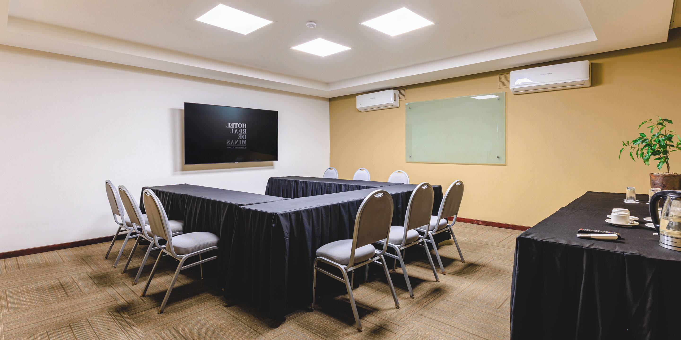 Een congreszaal met een lange tafel, stoelen, een projectorscherm en een airconditioning.