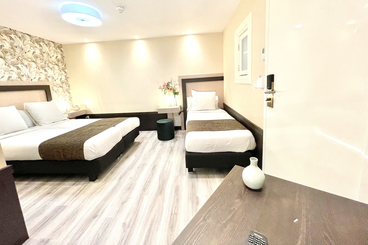 A hotel room with two beds, a small table, a vase, and a wall-mounted light fixture.