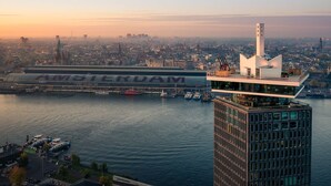 A modern high-rise building with a glass facade overlooking a river, with a large ship labeled "AMSTERDAM" docked in the background. - Hotel Oscar (Amsterdam)