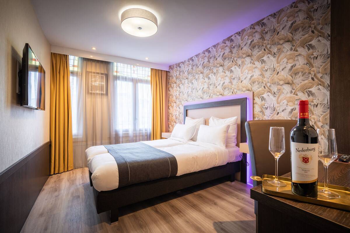 A hotel room with a bed, a TV mounted on the wall, a bottle of wine, and two glasses on a tray.