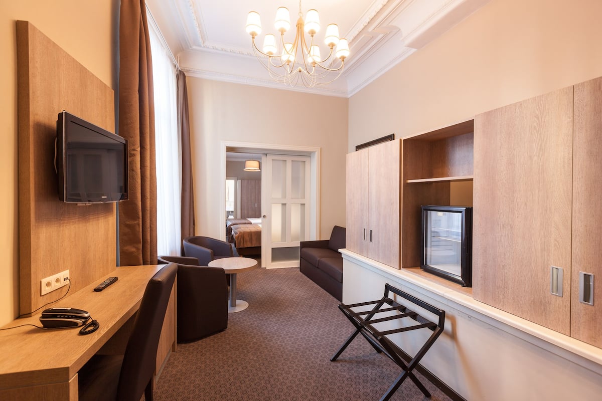 A hotel room with a TV mounted on the wall, a desk with a chair, a sofa, and a small table. There is a wardrobe with a mirror and a window.