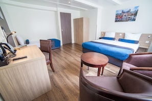 A modern hotel room with a wooden floor, a bed with blue bedding, a desk with a lamp, a chair, and a wardrobe. - Hotel Library Amsterdam (Amsterdam)