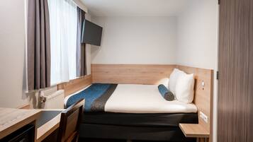 Une chambre d’hôtel de taille réduite, comprenant un lit, un bureau, une chaise, une télévision et une fenêtre avec des rideaux.