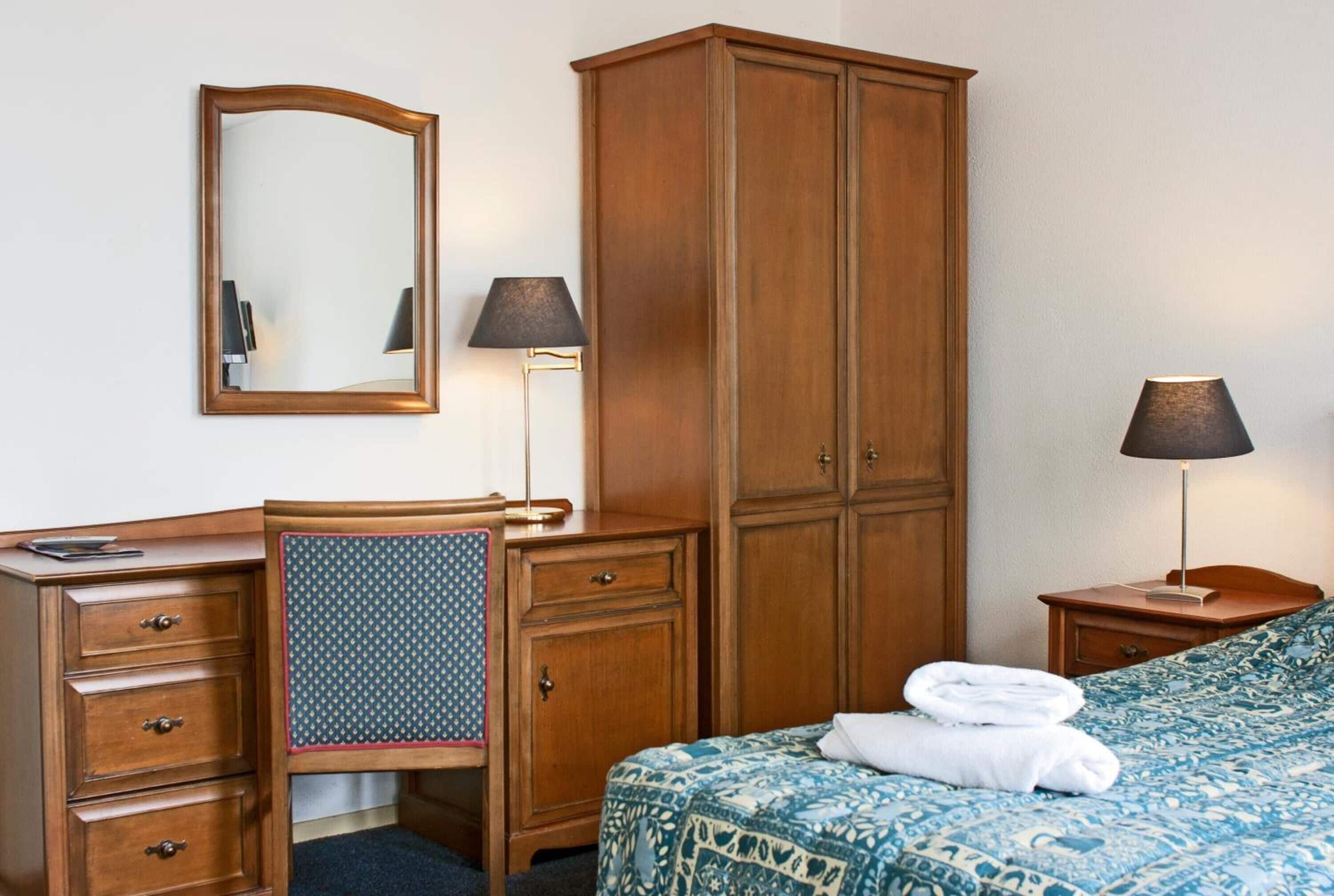 Een hotelkamer met een houten bureau, een grote kledingkast, een bed met een beddeken met dessin en twee bedlampjes.