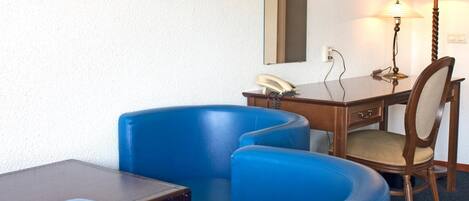 A hotel room with a desk, two blue armchairs, a wooden chair, a lamp, and a telephone.