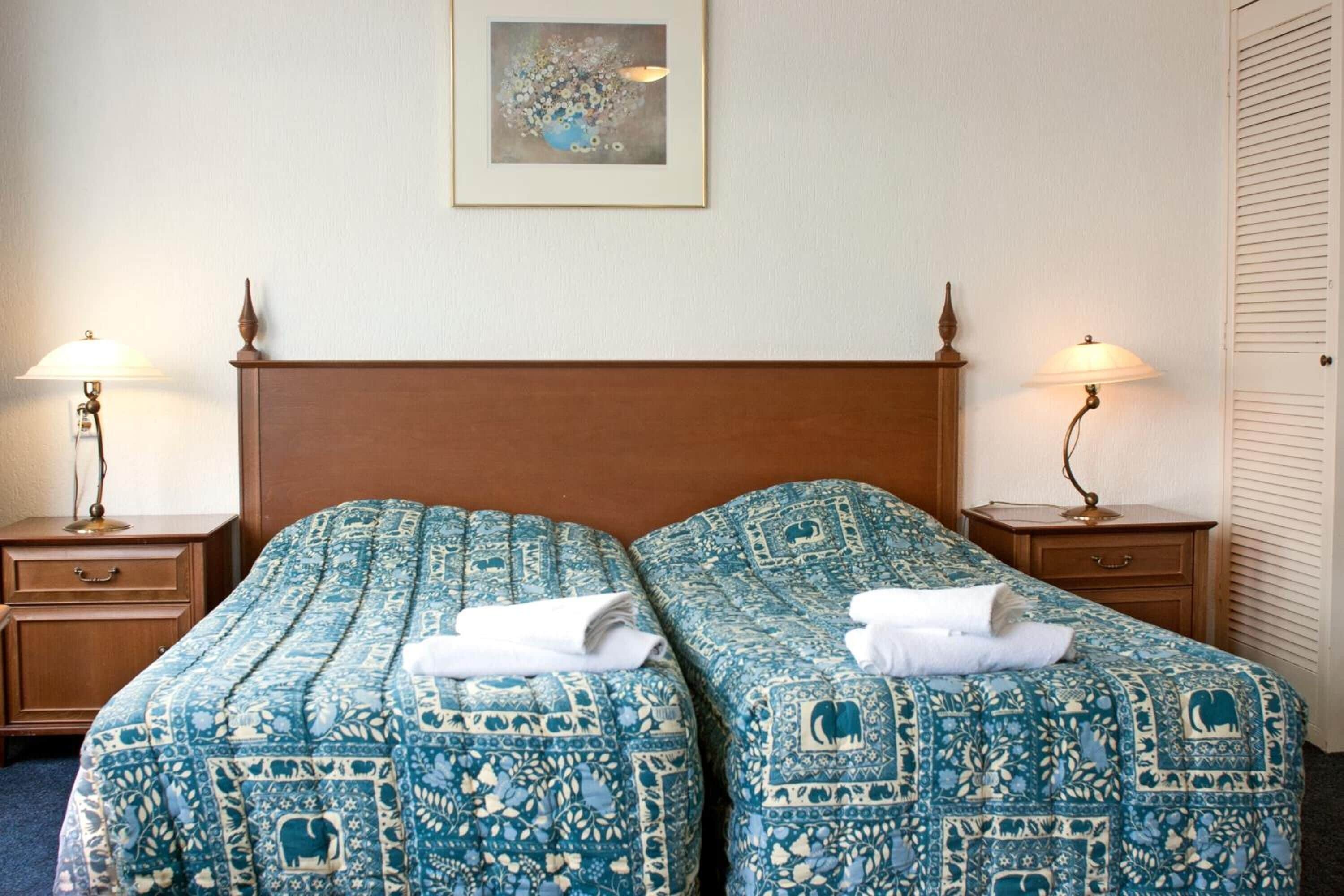 Een hotelkamer met twee bedden, elk voorzien van een blauw beddegoed met patroon, een houten hoofdbord, bedlampjes en een kunstwerk in een lijst aan de muur.