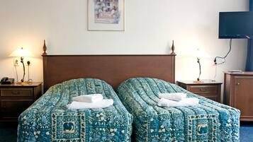 Een hotelkamer met twee bedden, elk voorzien van een blauw beddegoed met patroon, een houten hoofdbord, nachtkastjes met lampen, een televisie aan de muur en een ingelijst schilderij boven de bedden.