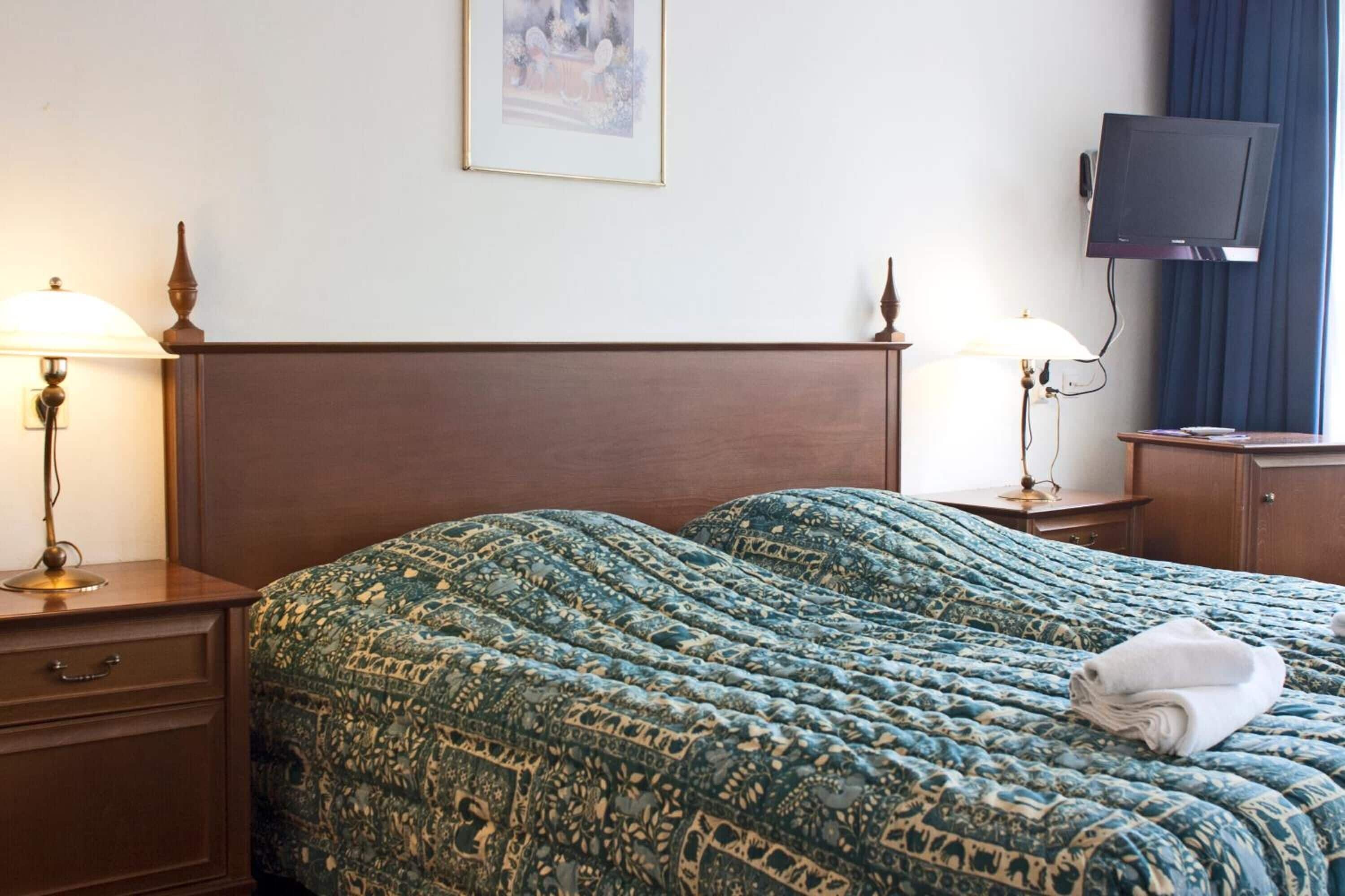 Ein Hotelzimmer mit einem großen Bett, zwei Nachttischen mit Lampen, einem an der Wand montierten Fernseher und einem gerahmten Bild über dem Bett.