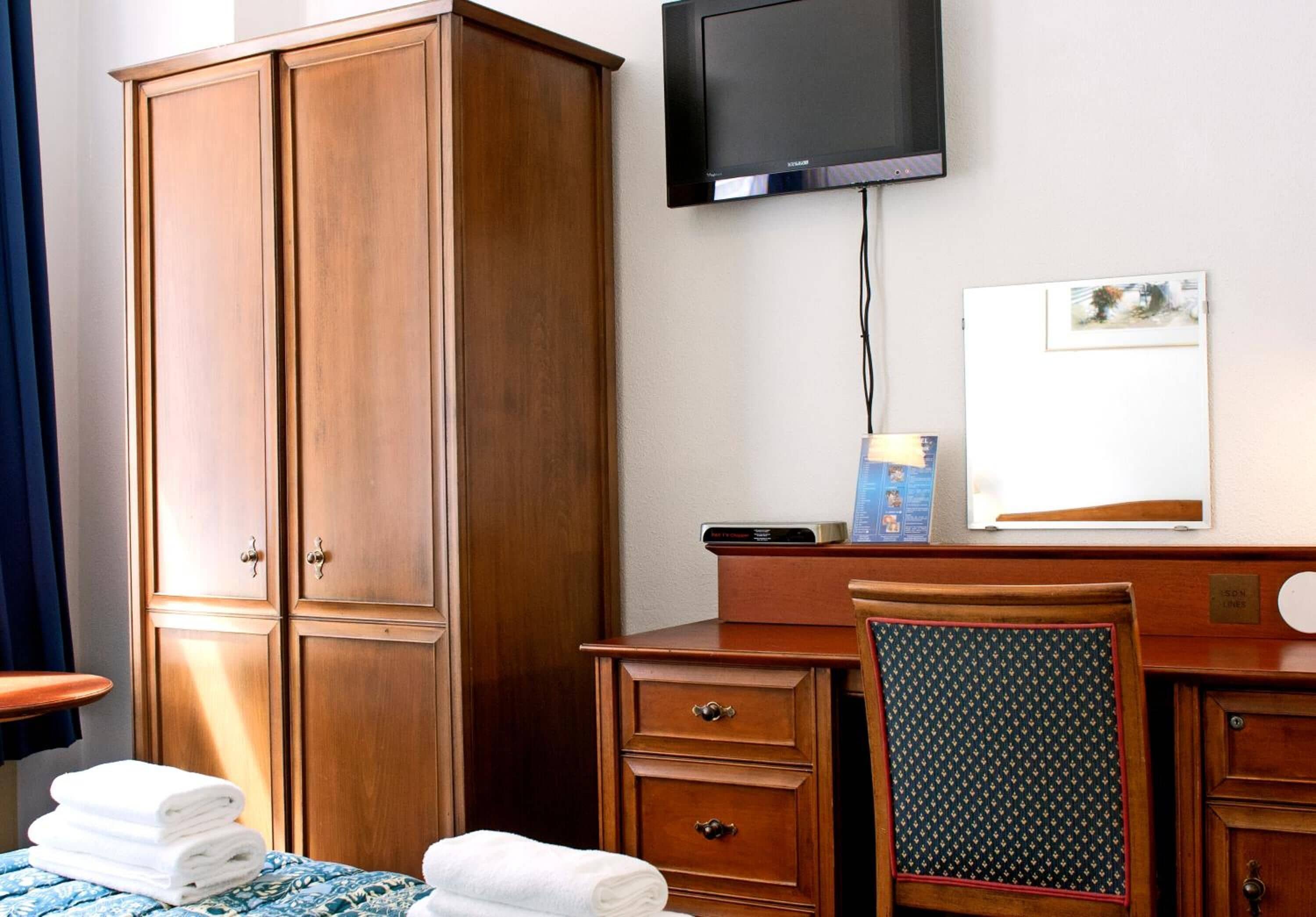 A hotel room with a wooden wardrobe, a desk with a chair, a television mounted on the wall, and a framed picture on the wall.