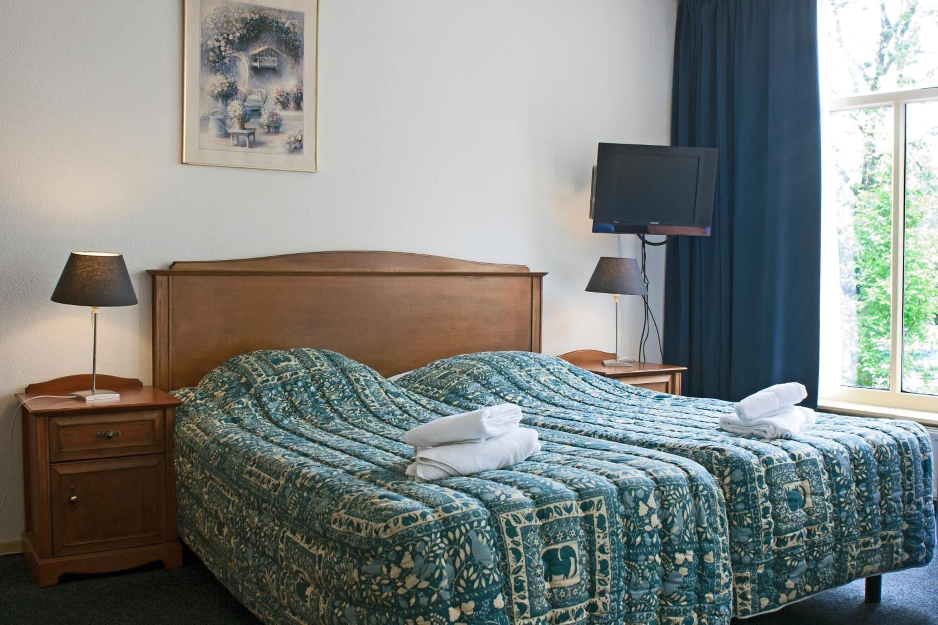 A hotel room with a large bed, two bedside tables, a TV, and a window with curtains.