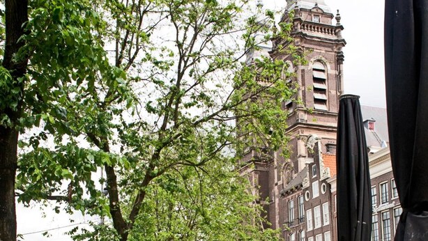 Ein Straßencafé mit Korbstühlen und Tischen, im Hintergrund ein historischer Glockenturm und rote Blumen in Pflanzkästen.
