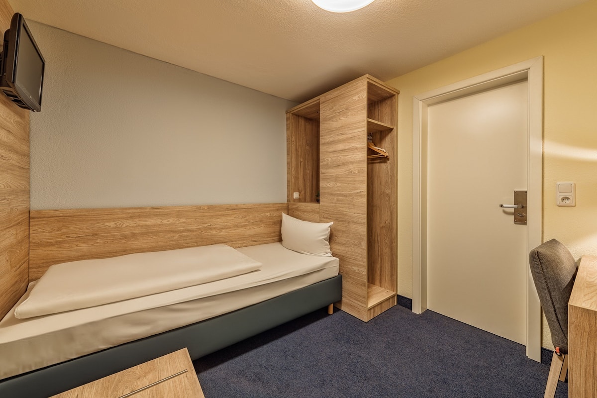 Ein kleiner, ordentlicher Raum mit einem Einzelbett, einem Holzschrank, einem Schreibtisch und einem Stuhl.