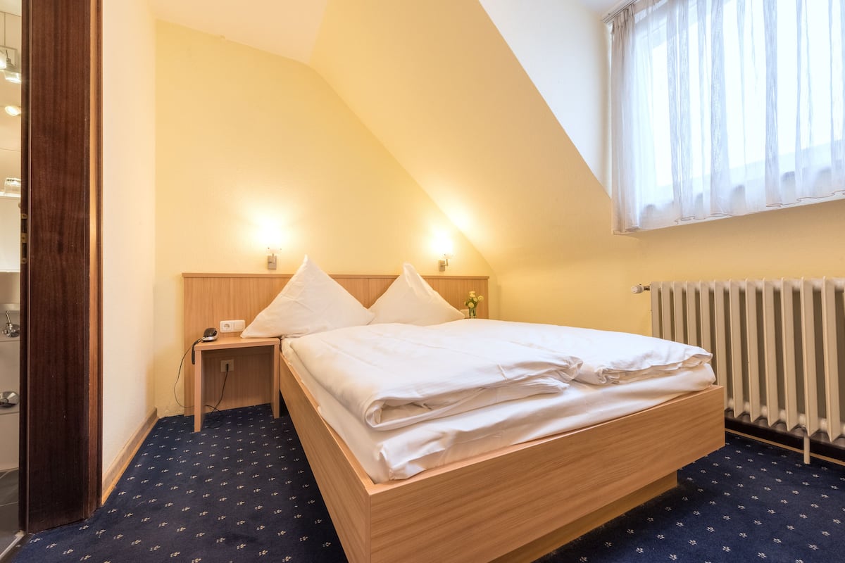 Ein Schlafzimmer mit einem hölzernen Bett, einem Nachttisch, einem Beistelltisch und einem Fenster mit durchsichtigen Vorhängen.