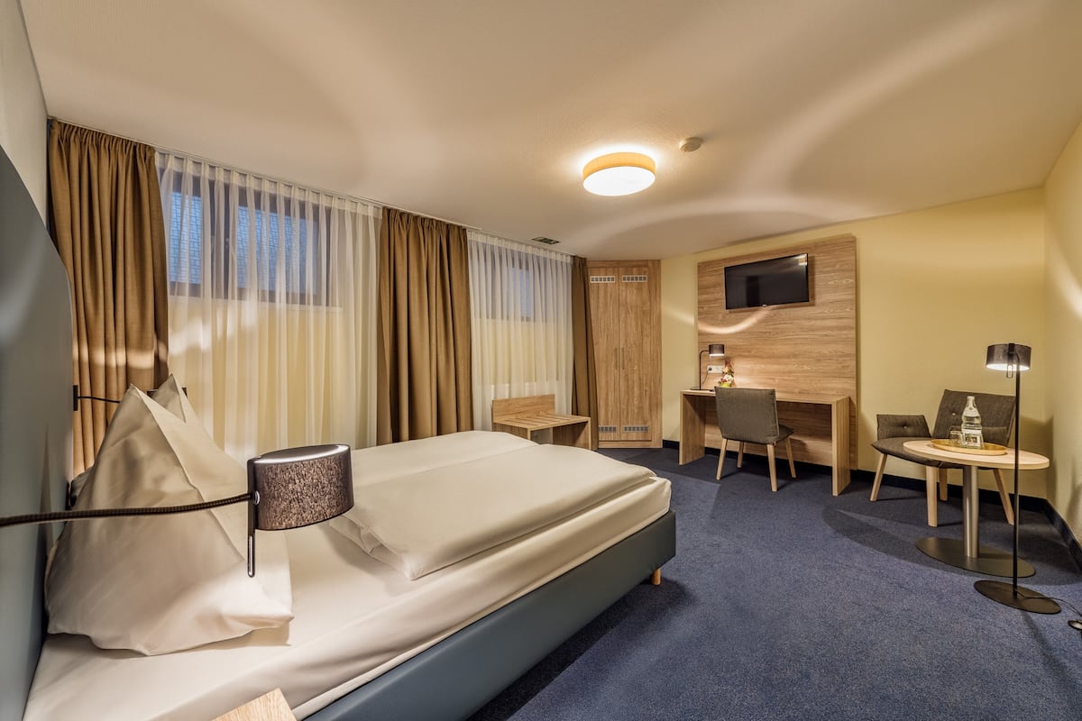 Ein Hotelzimmer mit einem Bett, einem Schreibtisch, einem Stuhl, einem an der Wand befestigten Fernseher und einem kleinen Tisch mit einer Lampe.
