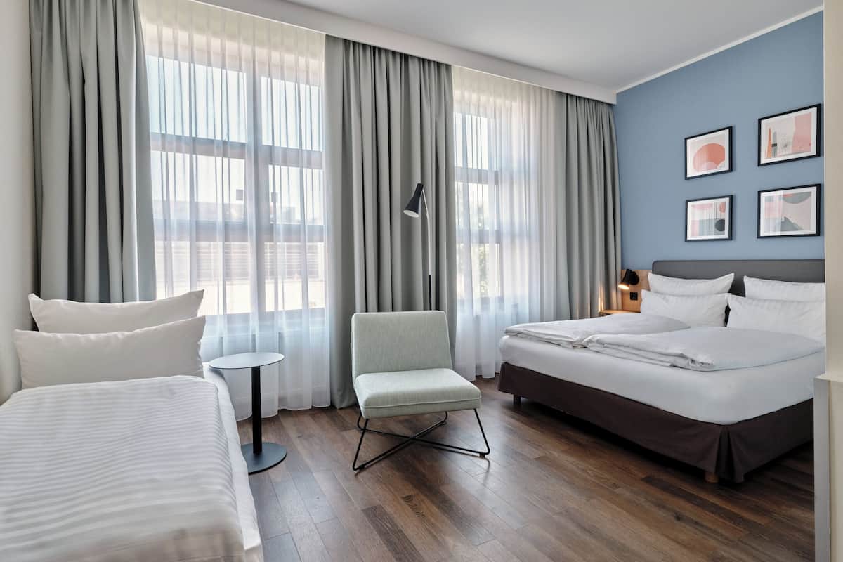 Habitación de hotel con una cama grande, una cama más pequeña, una silla, una mesa redonda y vistas al exterior a través de cortinas traslúcidas.