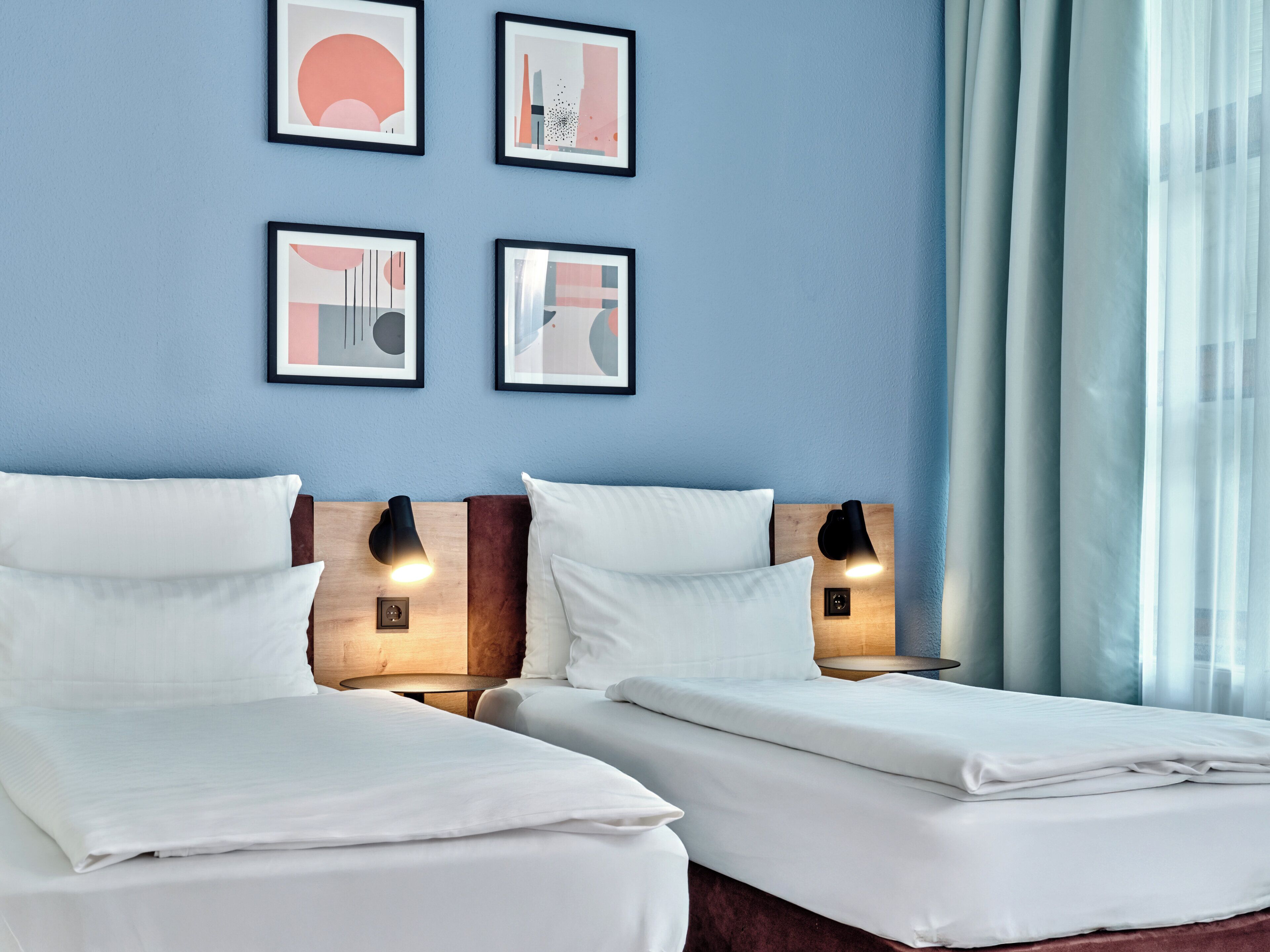 A hotel room with two beds, a wooden headboard, a nightstand with a lamp, and framed artwork on the wall.