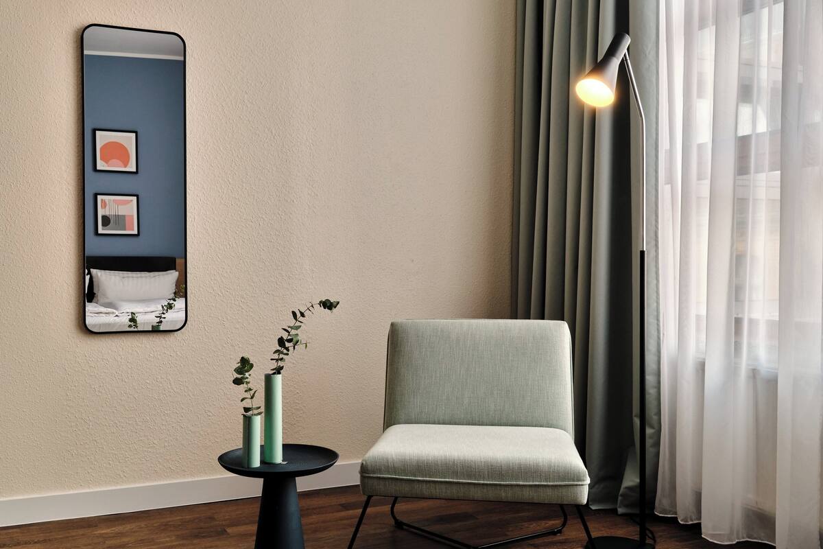 Una habitación moderna con un sillón gris, una mesa redonda con un florero, un espejo y una lámpara de pie.
