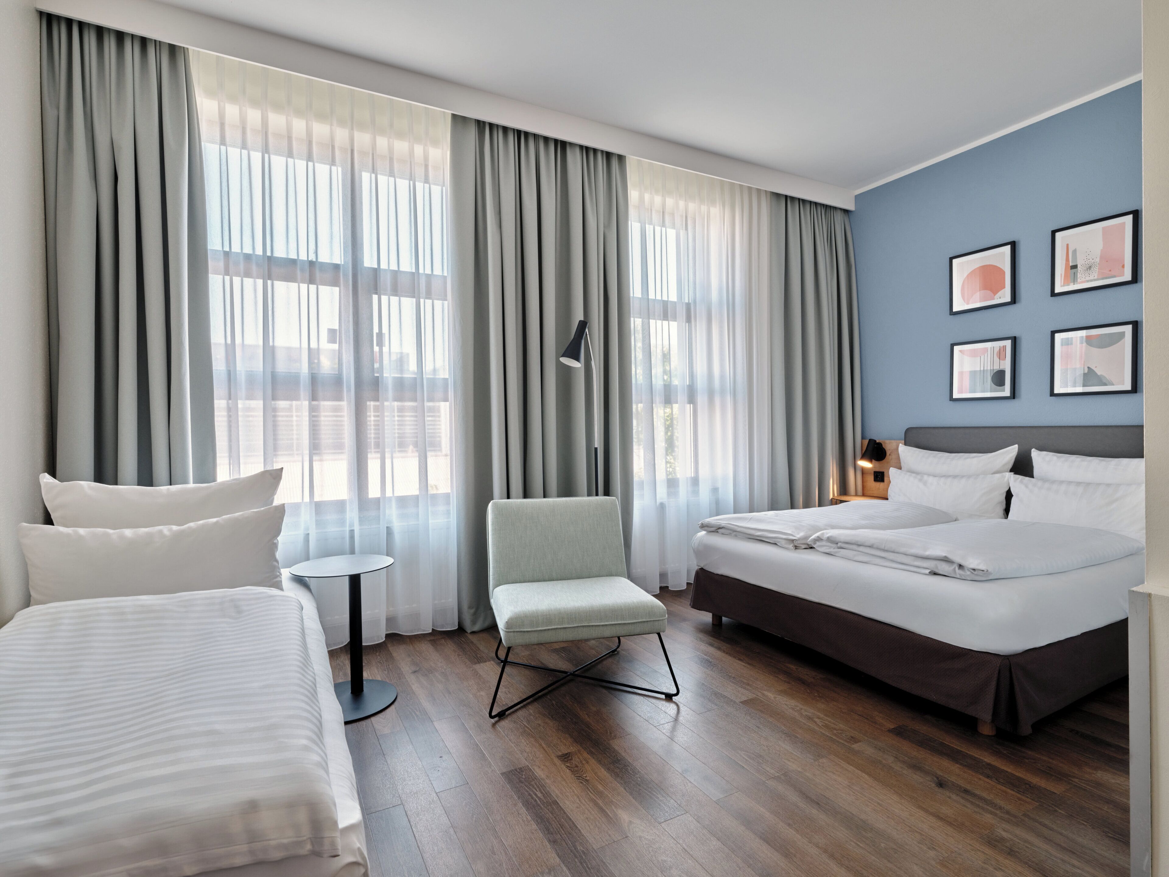 A hotel room with a large bed, a smaller bed, a chair, a round table, and a view of the outdoors through sheer curtains.