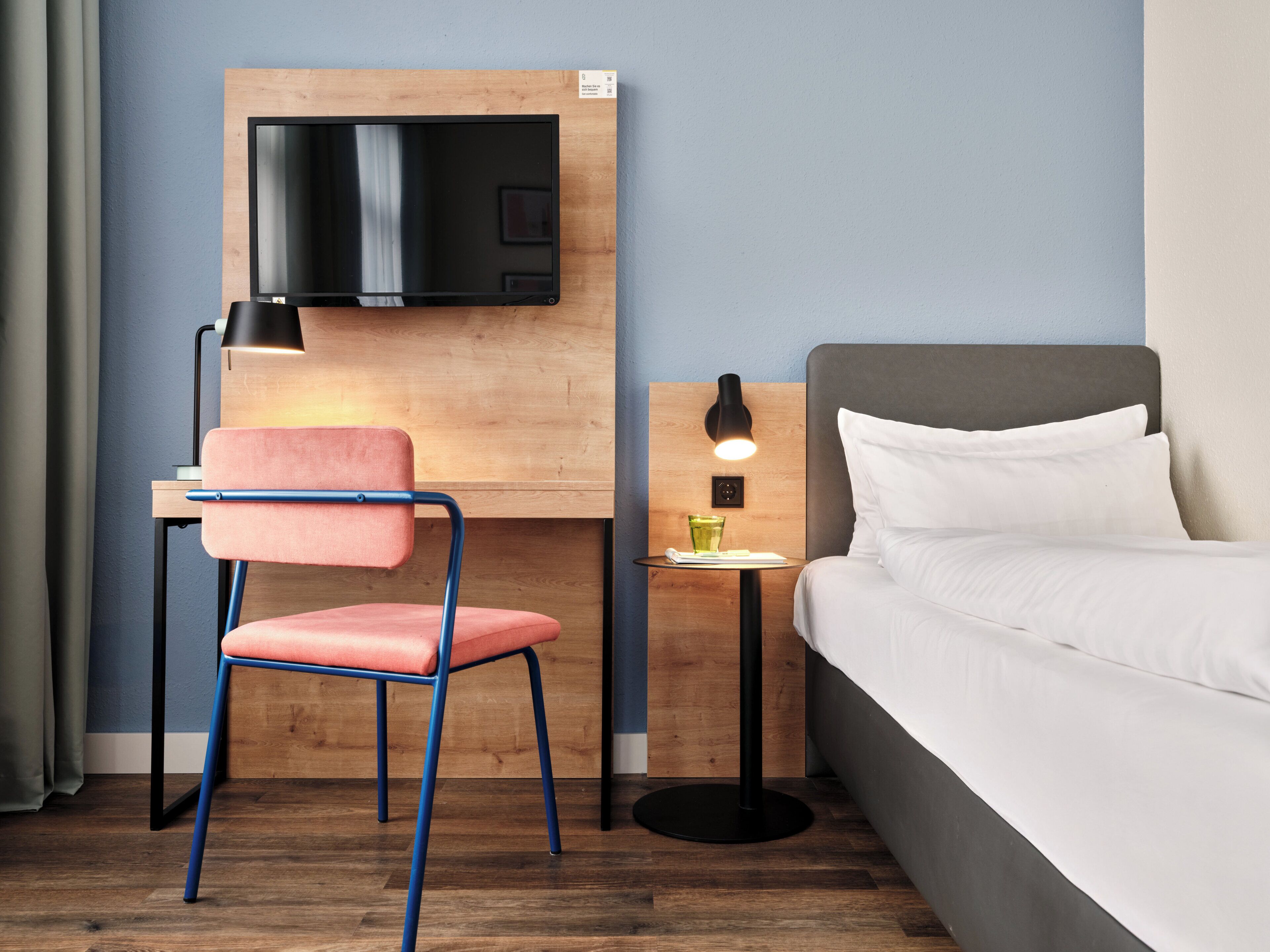 A hotel room with a bed, a desk with a TV, a chair, a bedside table with a lamp, and a small table with a glass.