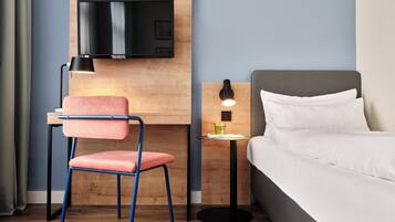 A hotel room with a bed, a desk with a TV, a chair, a bedside table with a lamp, and a small table with a glass.