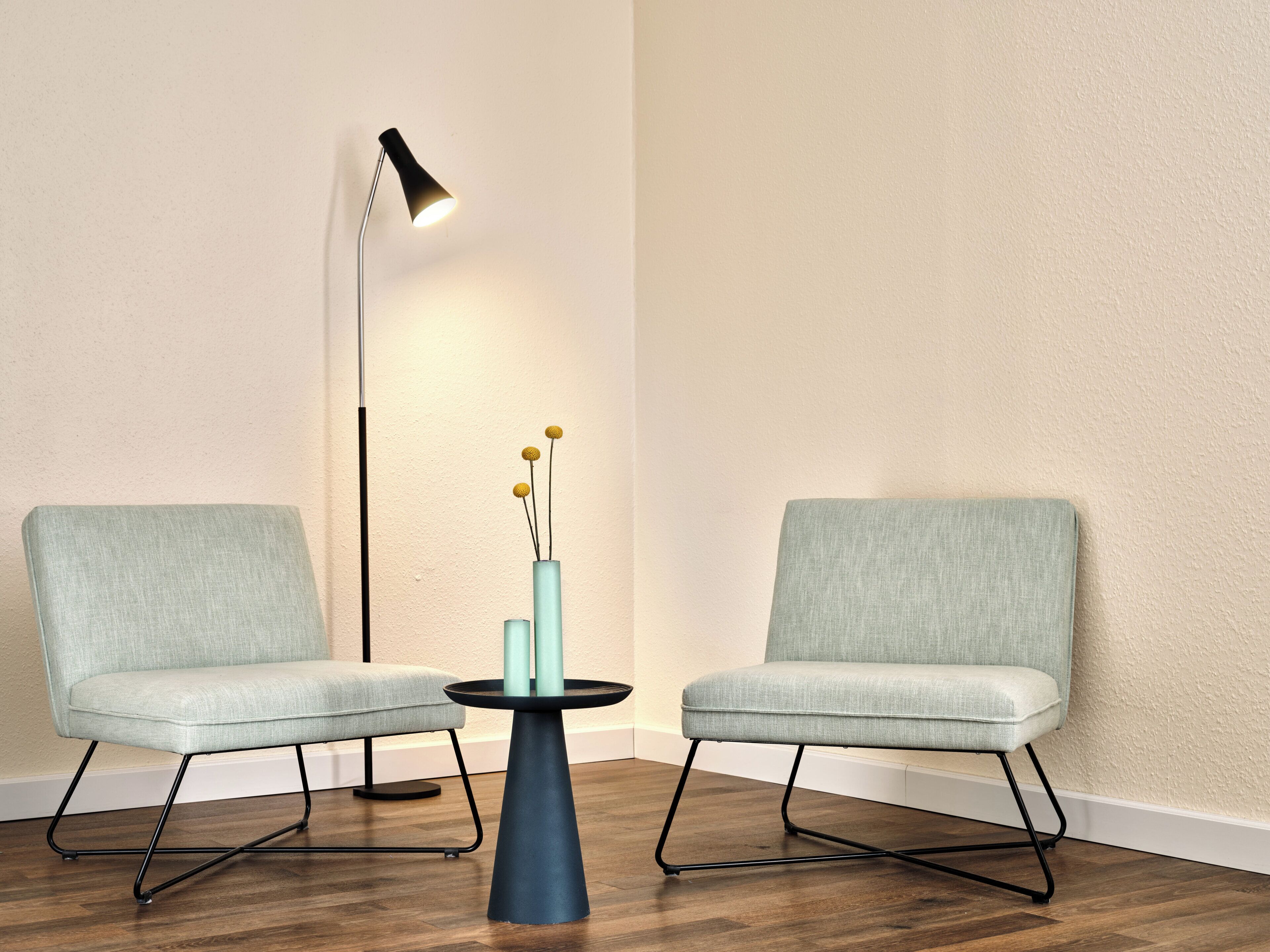Dos sillas, una mesa y una lámpara de pie en una habitación con suelo de madera y una pared lisa.