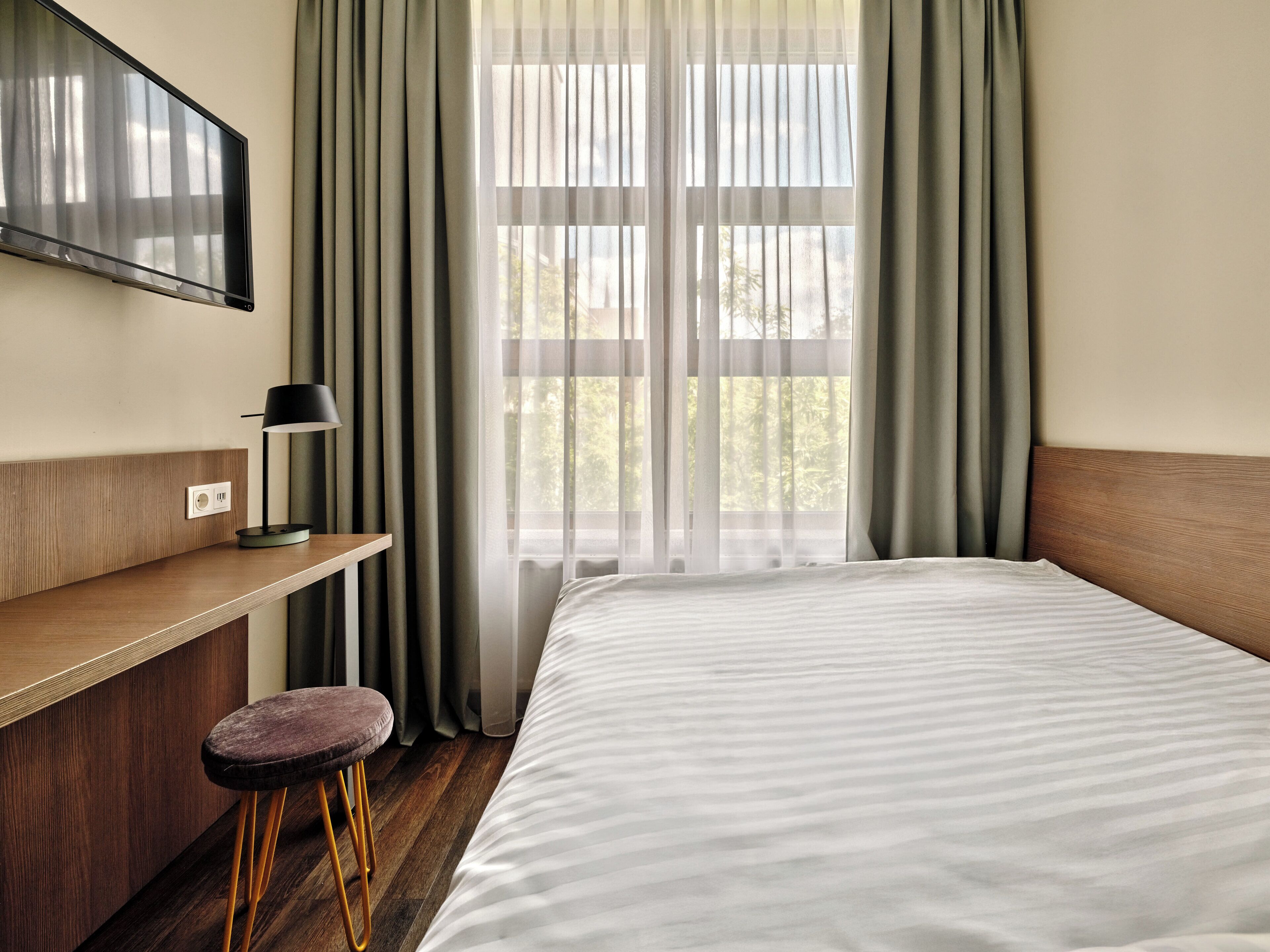 Habitación de hotel con cama, escritorio, silla, televisor en la pared y ventana con cortinas.
