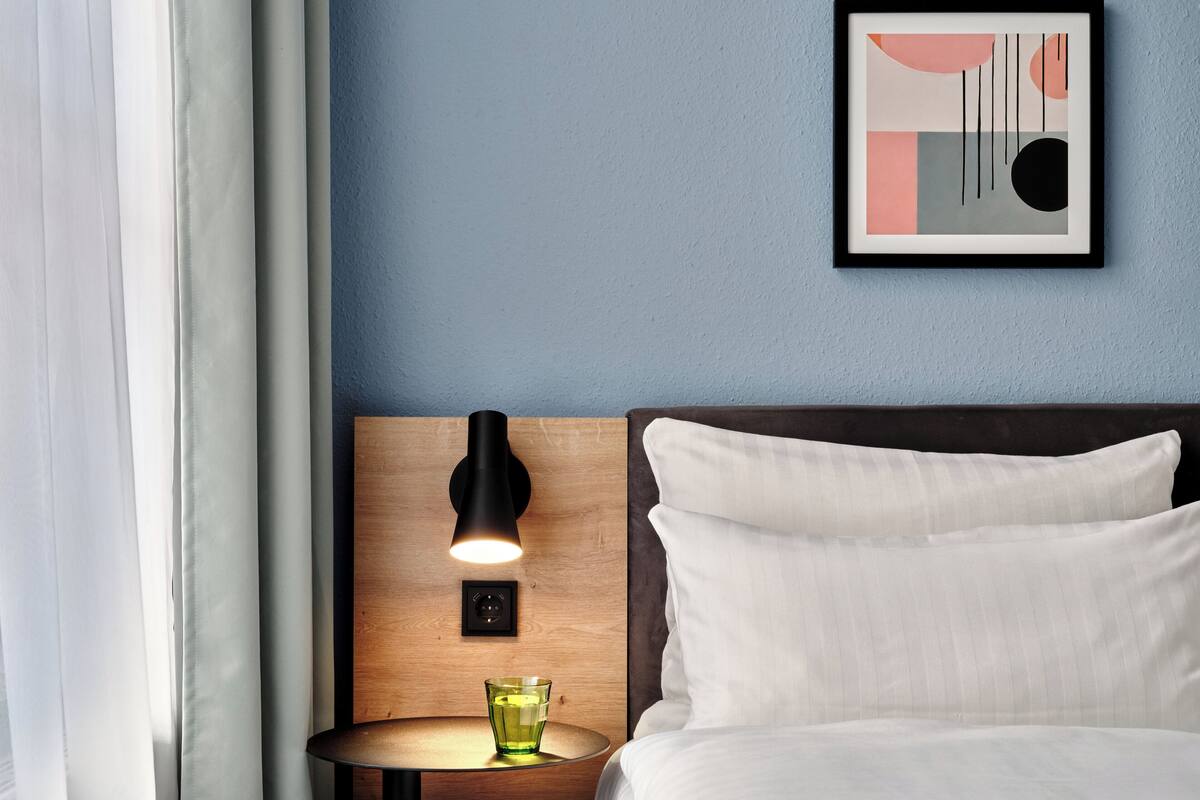 Una cama bien hecha con ropa blanca, un cabecero de madera, una mesita de noche con una lámpara negra y una obra de arte abstracta enmarcada en la pared.