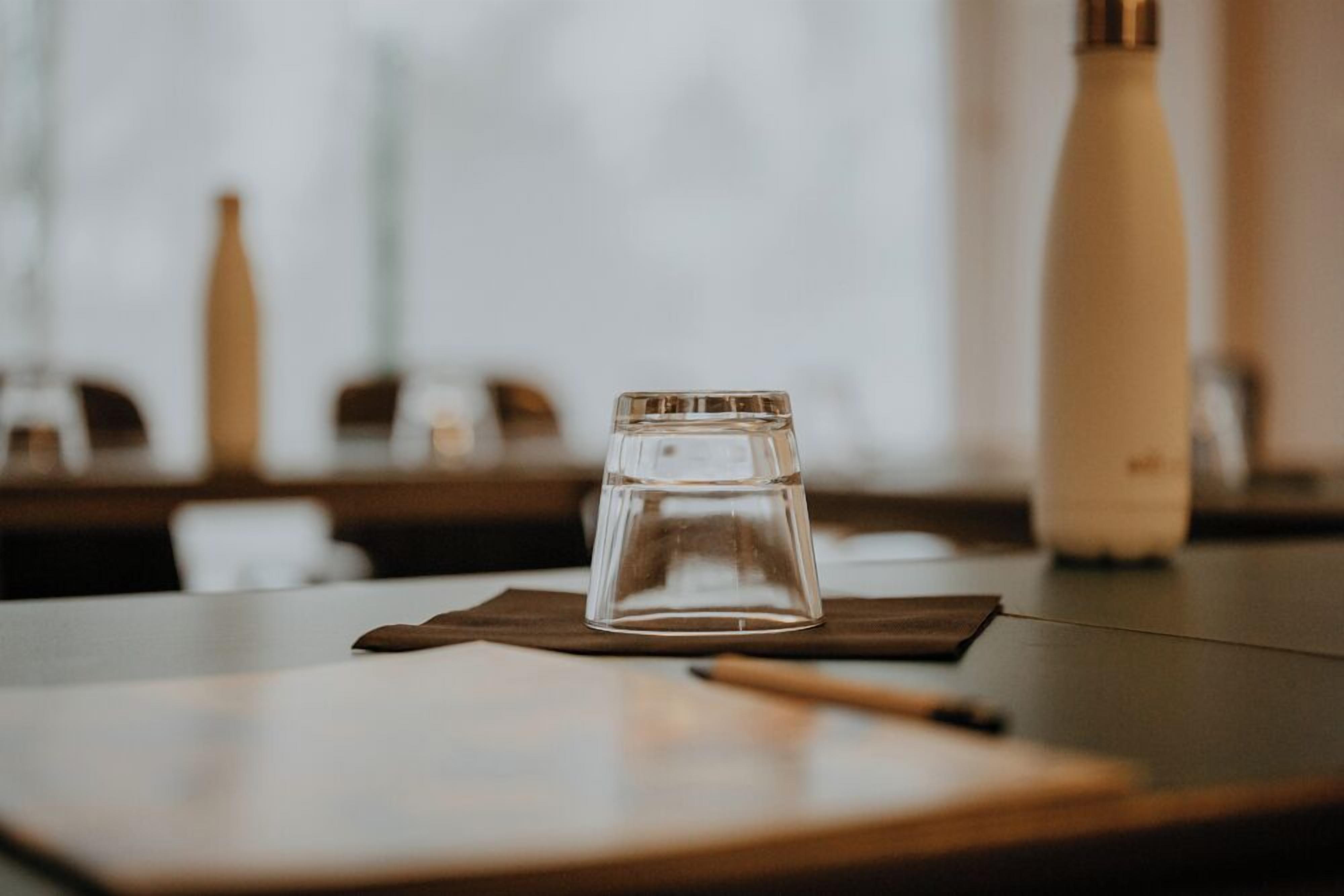 A clear glass tumbler on a wooden table with a napkin underneath, a pen beside it, and a white bottle in the background.