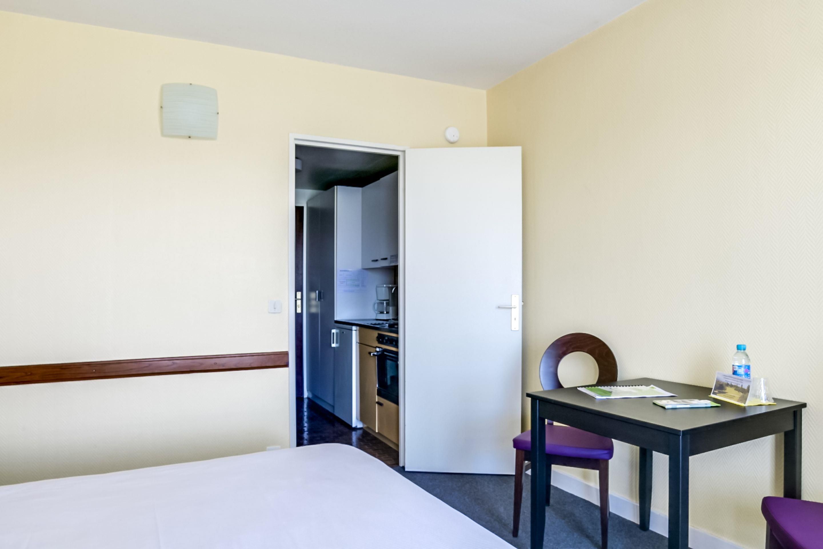 A hotel room with a bed, a table with a chair, a small table with a chair, a water bottle, and a glass.