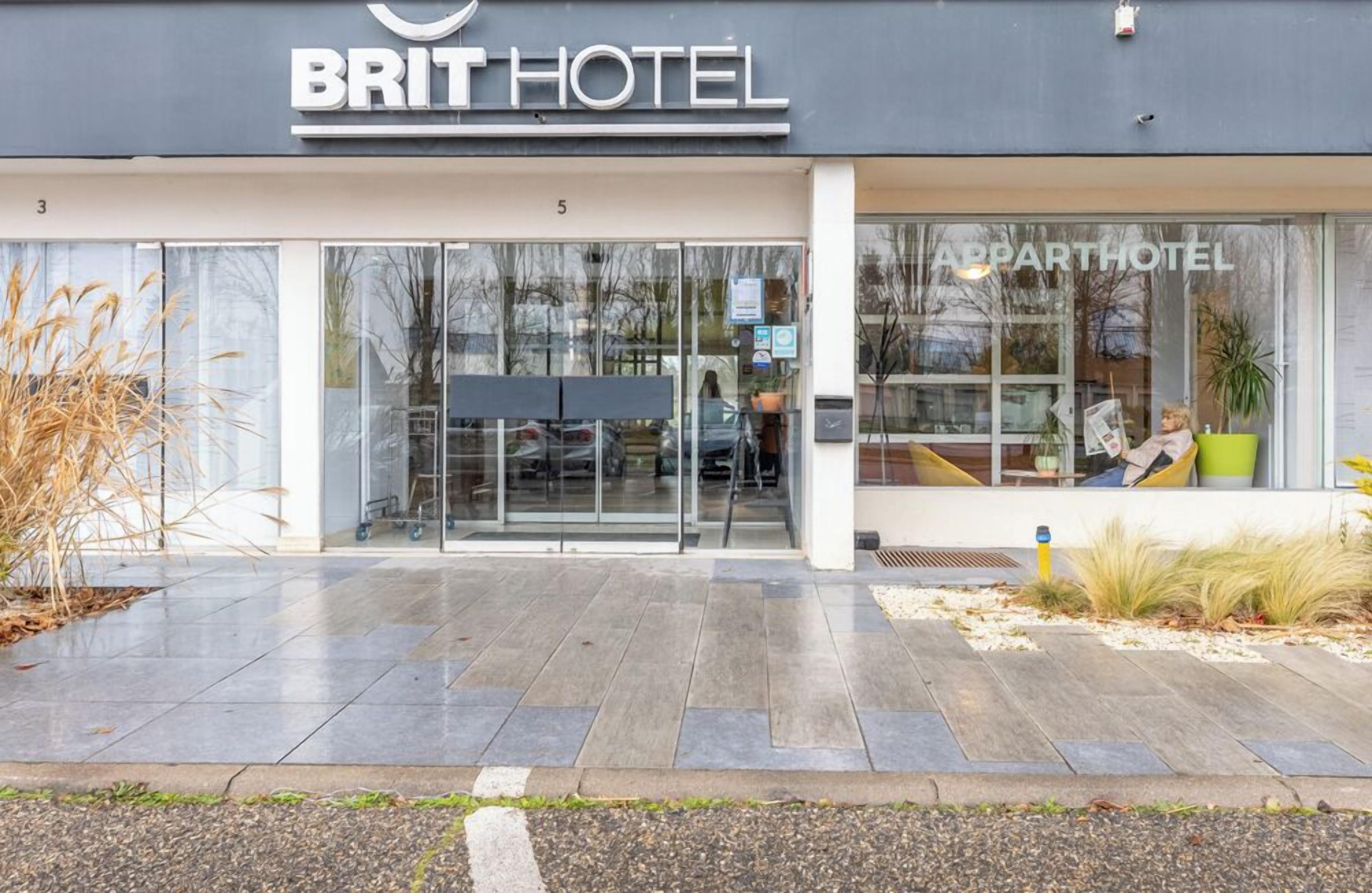 L’entrée d’un hôtel de la chaîne BRIT HOTEL, avec une façade en verre, un espace extérieur aménagé avec des sièges, et une enseigne indiquant qu’il s’agit d’un hôtel proposant des appartements.
