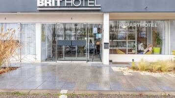 L’entrée d’un hôtel de la chaîne BRIT HOTEL, avec une façade en verre, un espace extérieur aménagé avec des sièges, et une enseigne indiquant qu’il s’agit d’un hôtel proposant des appartements.