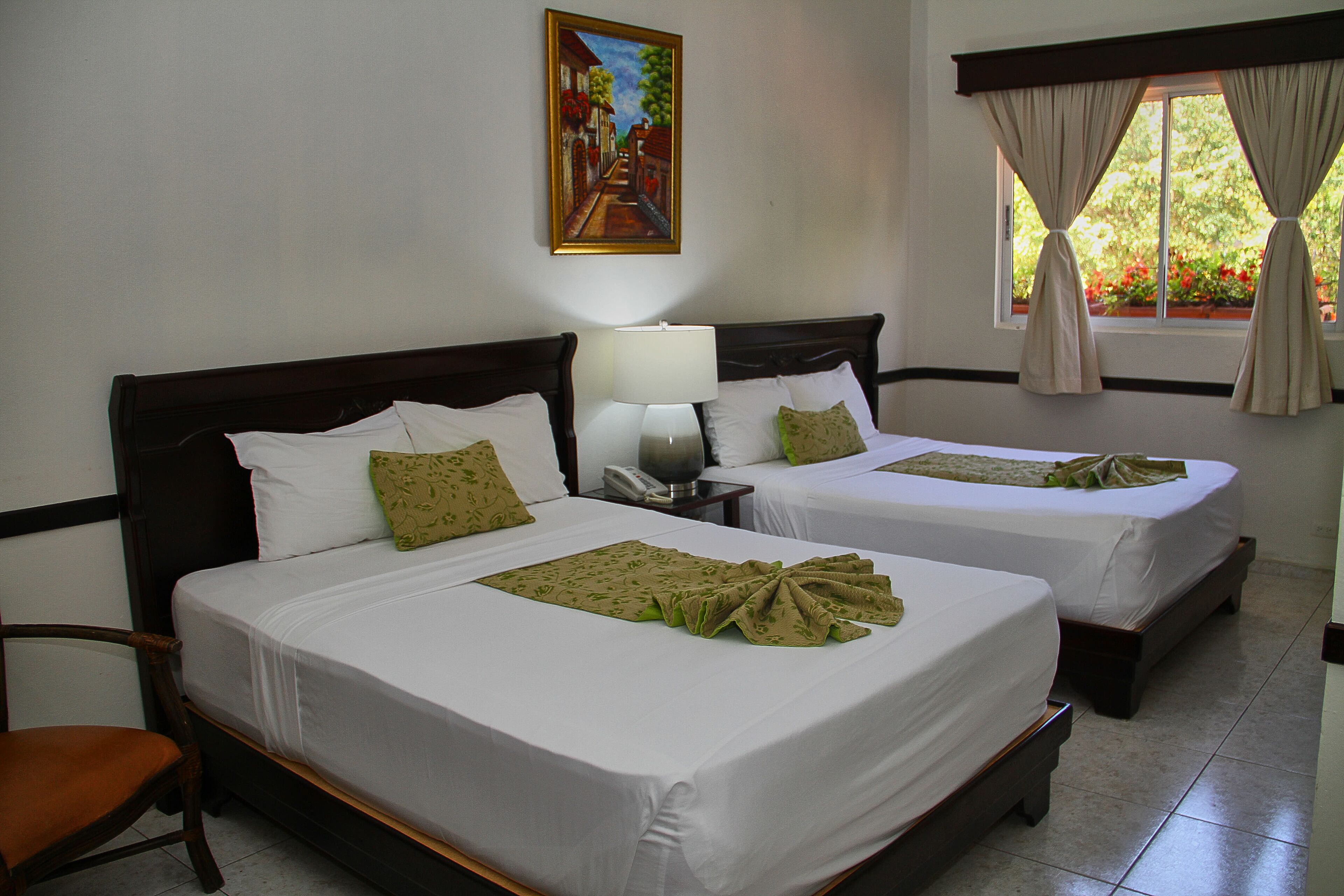 A hotel room with two beds, a nightstand with a lamp, a painting on the wall, and a window with curtains.