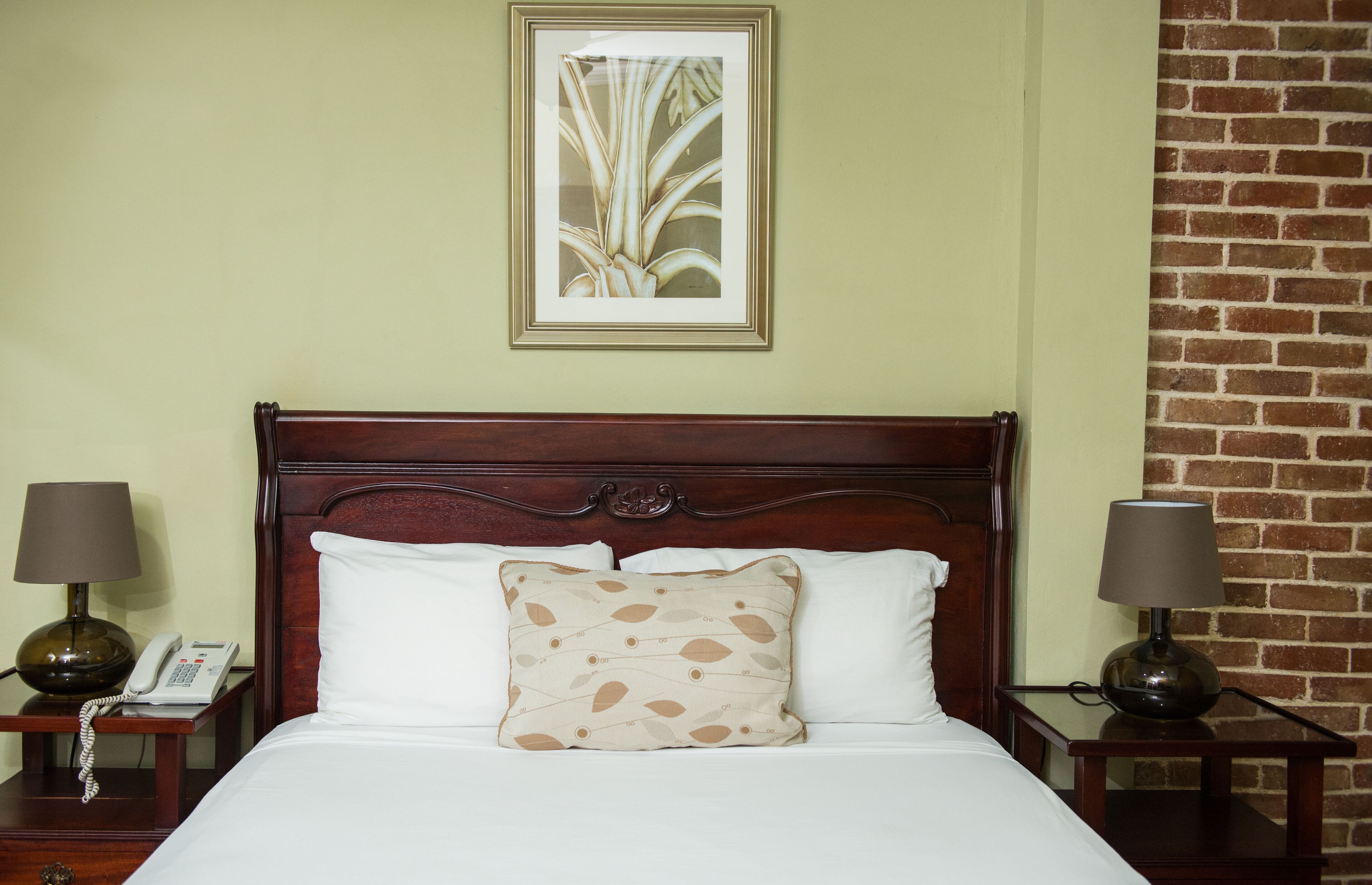 A hotel room with a wooden headboard, a bed with white linens, a patterned pillow, two bedside lamps, and a framed artwork on the wall.
