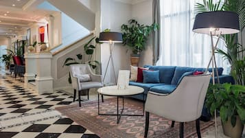 Una hall d'albergo con pavimento a scacchi, un divano blu, una poltrona bianca, un tavolino rotondo e una lampada da terra.