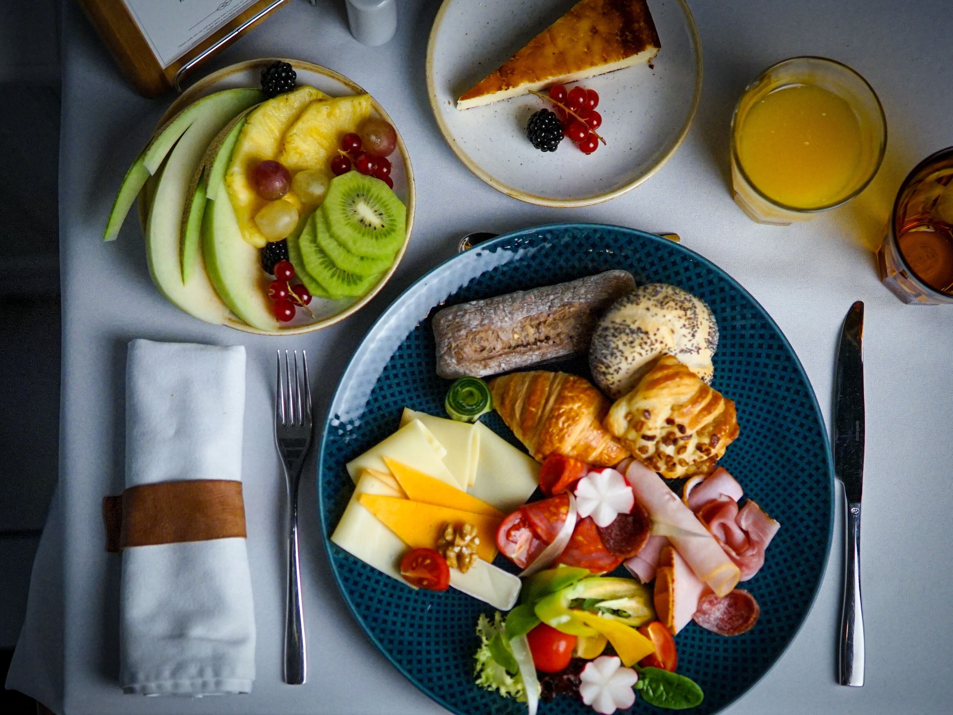 Une table dressée pour le petit-déjeuner, avec une assiette garnie d’un assortiment de fruits, de fromage, de charcuterie et de viennoiseries, accompagnée d’un verre de jus d’orange et d’une part de tarte.