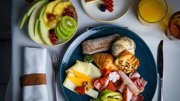 Une table dressée pour le petit-déjeuner, avec une assiette garnie d’un assortiment de fruits, de fromage, de charcuterie et de viennoiseries, accompagnée d’un verre de jus d’orange et d’une part de tarte.