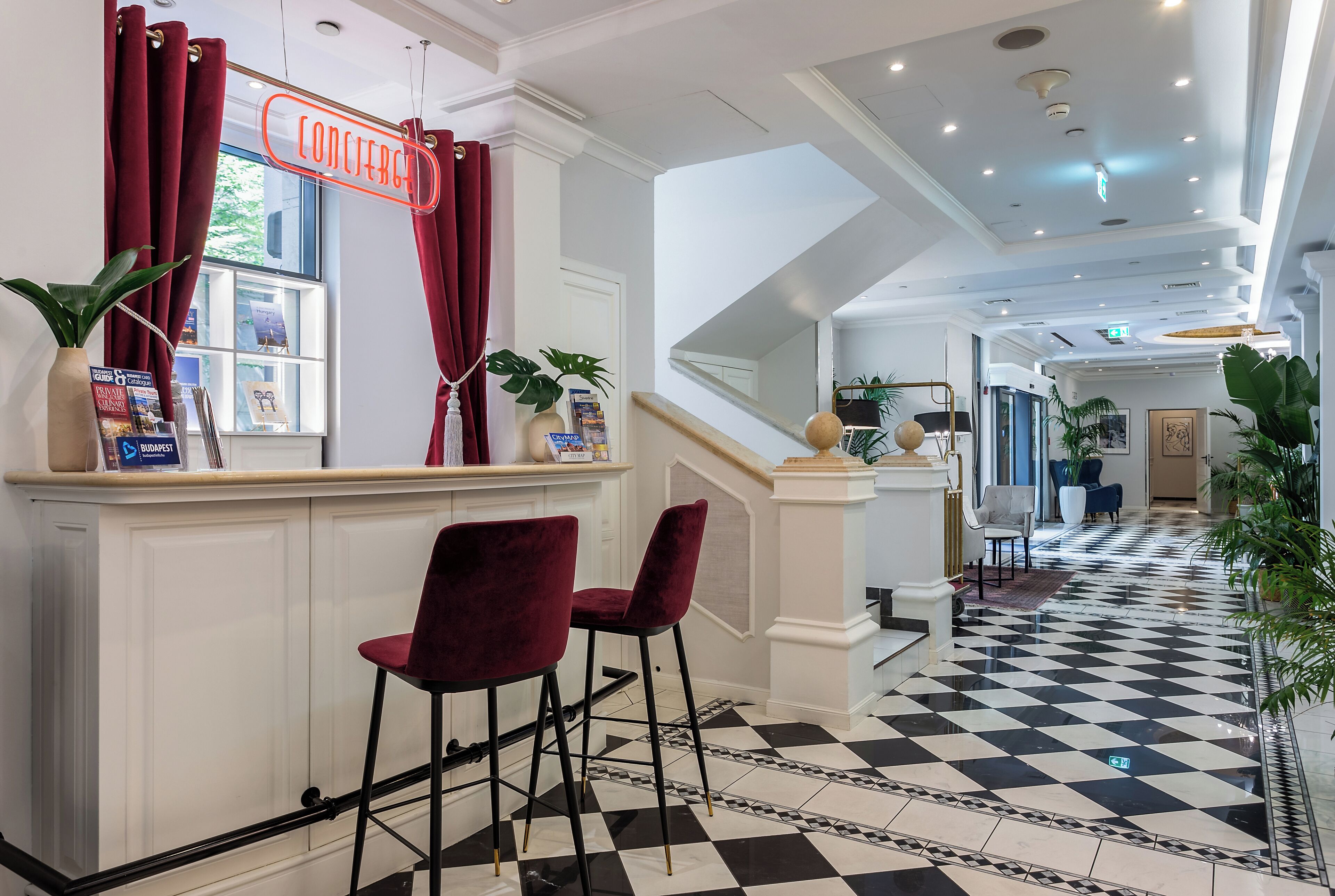 Un hall d’hôtel avec un comptoir d’enregistrement, des fauteuils en velours rouge, un sol à carreaux noirs et blancs et un escalier menant à d’autres espaces.