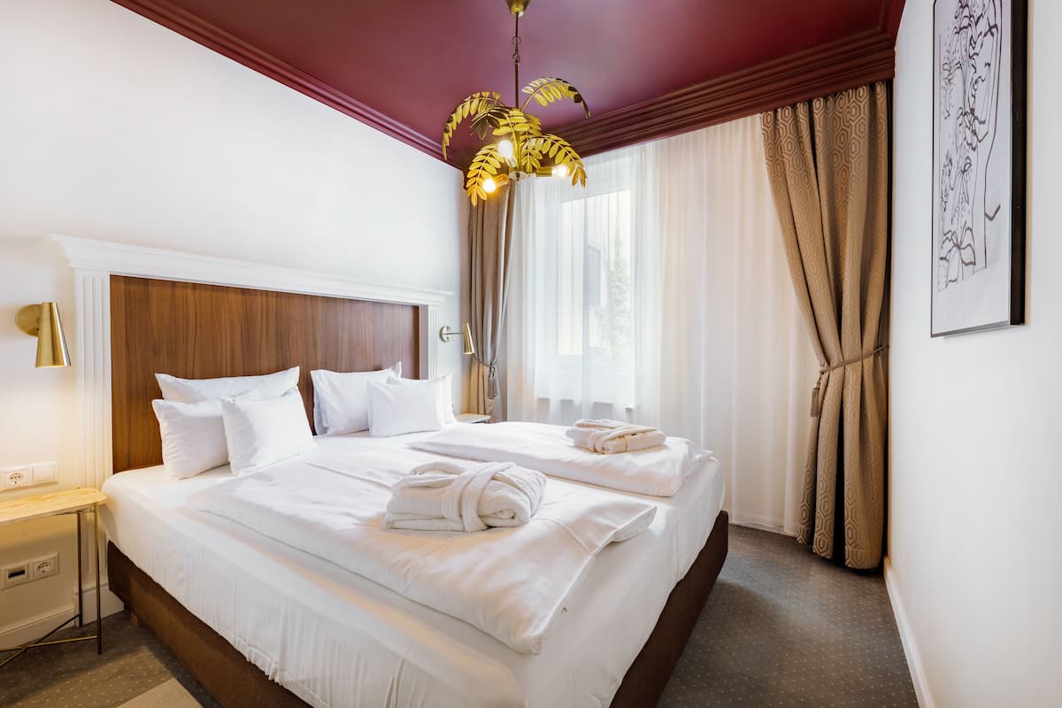 A hotel room with two beds, a wooden headboard, a bedside table with a lamp, and a large window with patterned curtains.