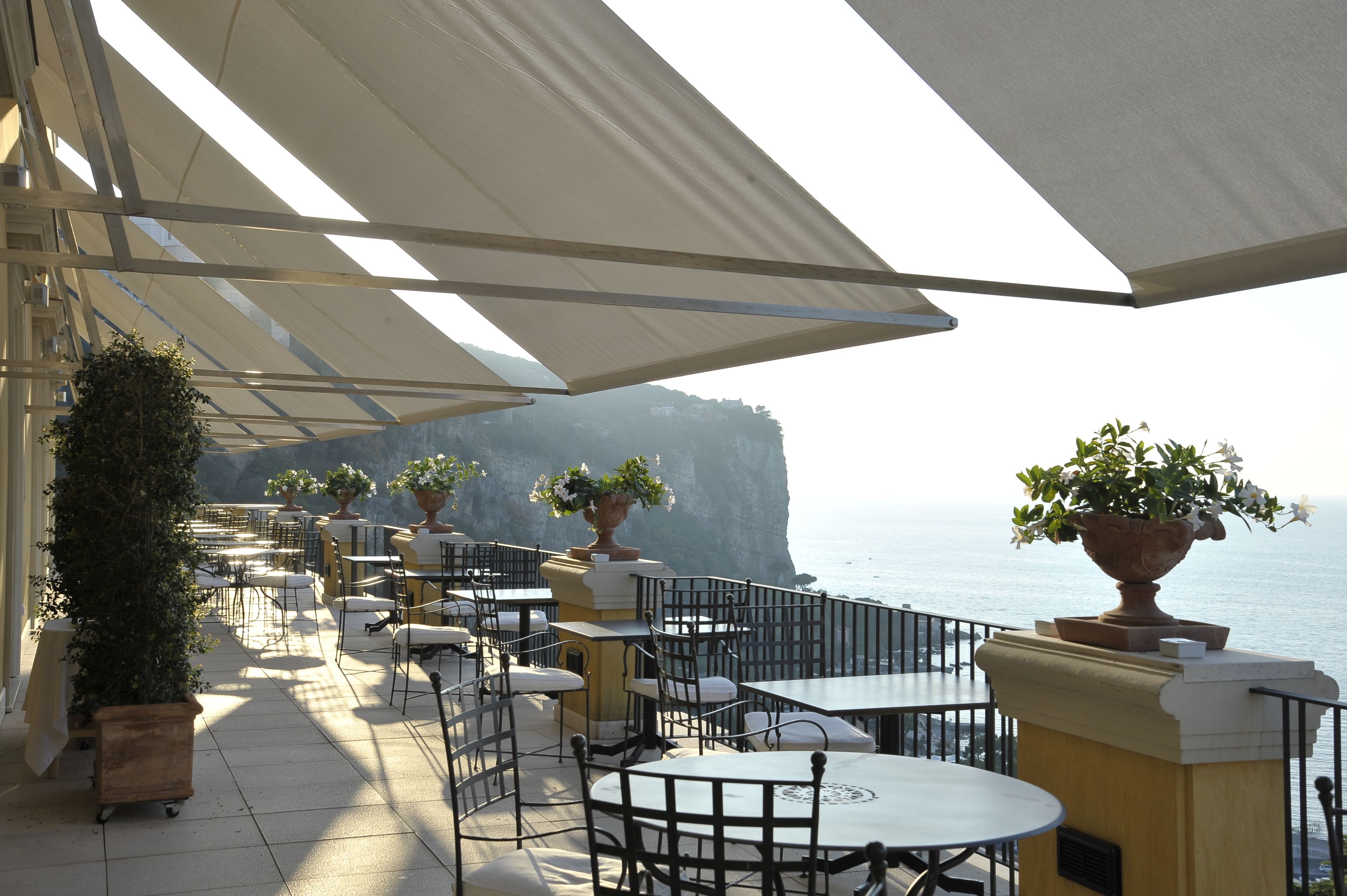 An outdoor terrace with tables and chairs, potted plants, and a view of the sea.