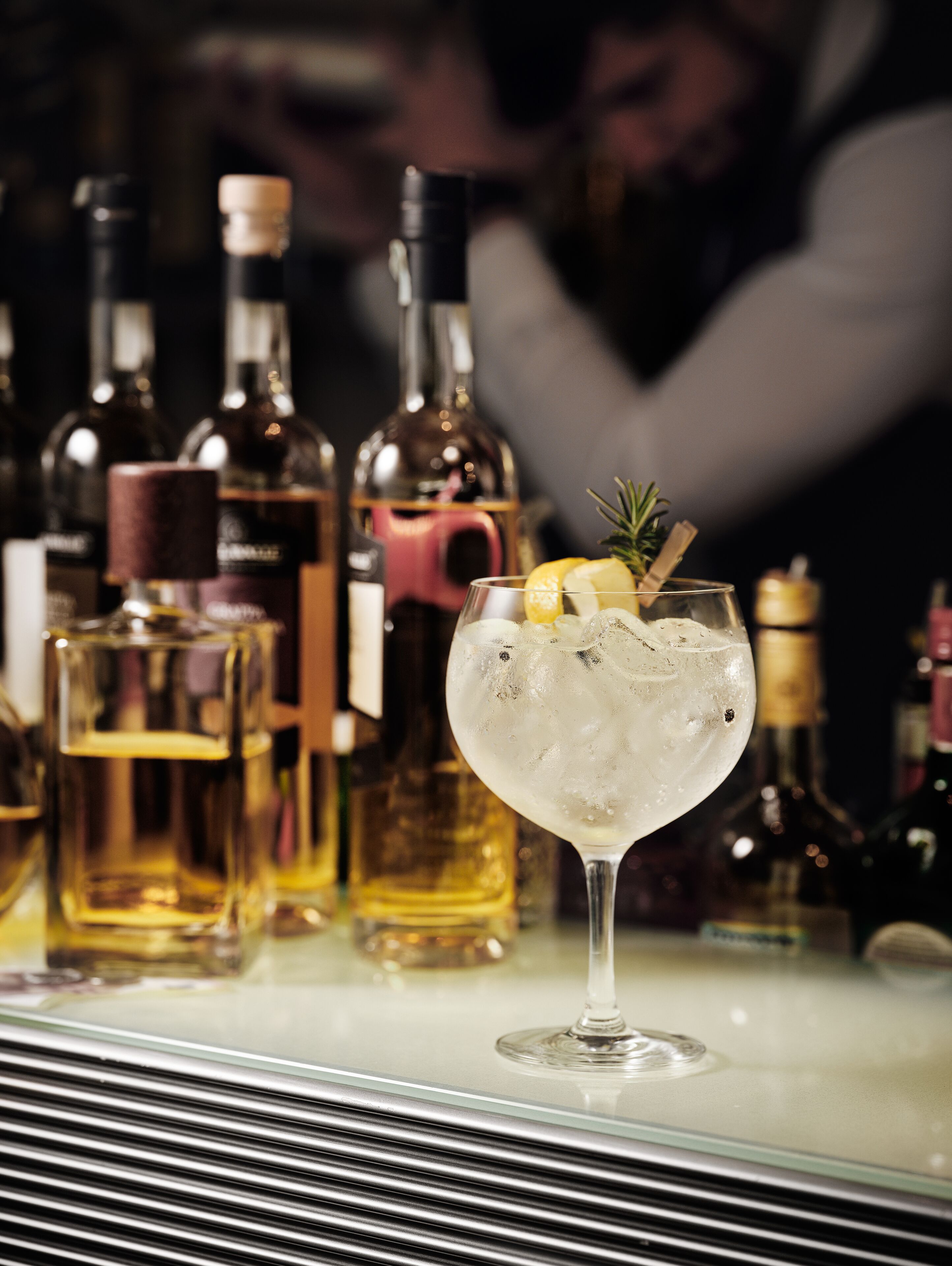 Un bar con una copa de cóctel adornada con una rodaja de limón y una ramita de romero, rodeada de diversas botellas de alcohol.