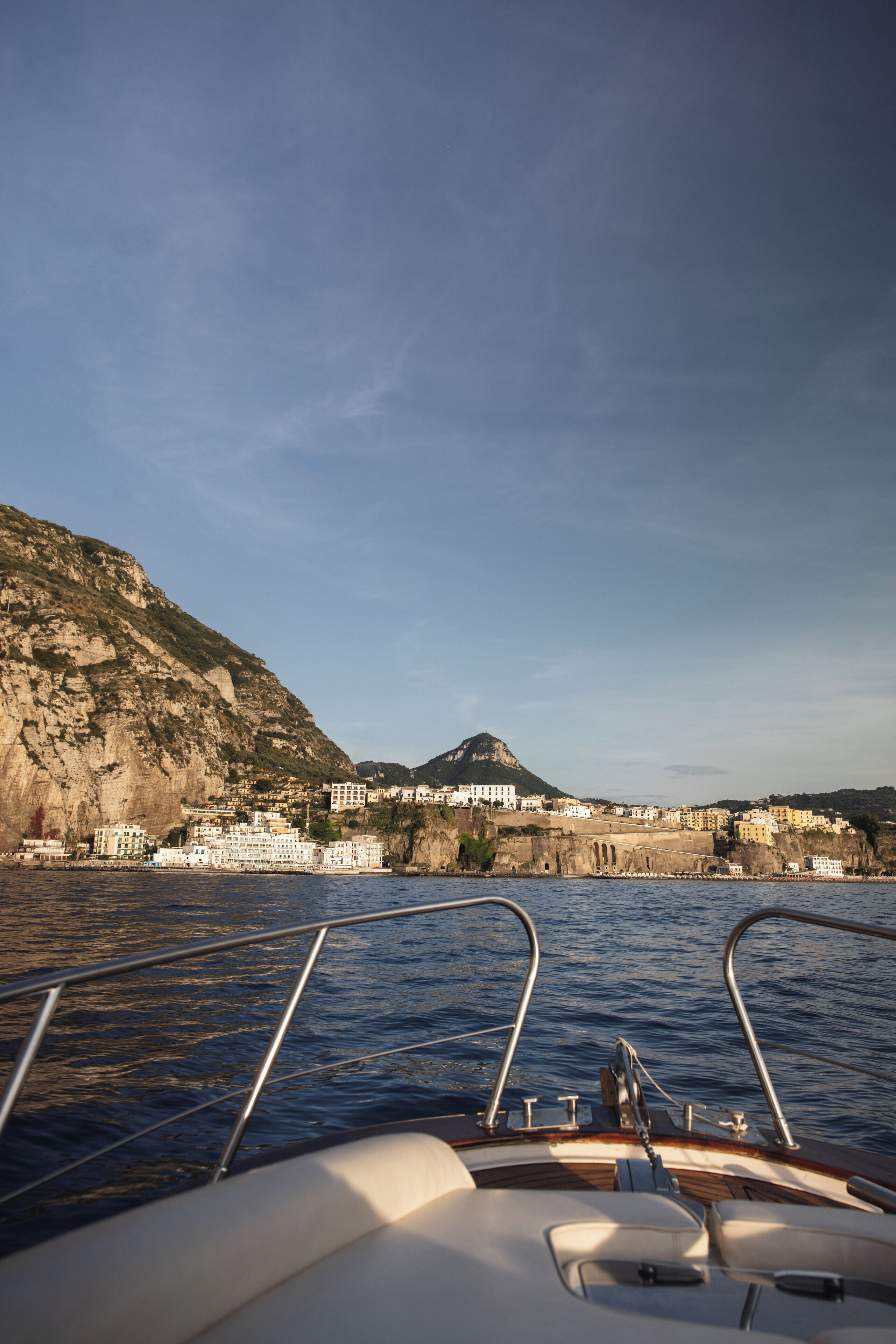 Convés de barco com grades de metal e vista para uma vila costeira, com edifícios e uma montanha ao fundo.