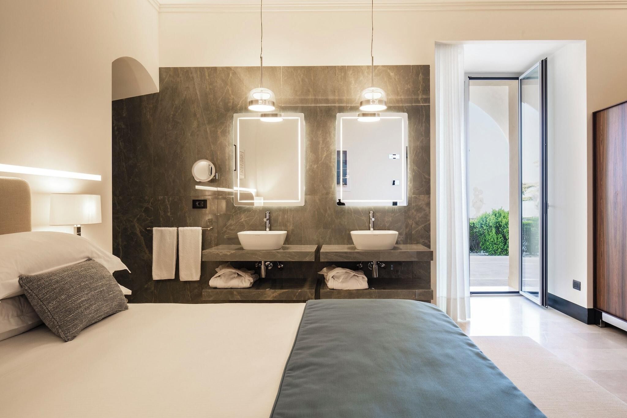 A modern hotel room with a large bed, two bedside tables, a double sink, and a large mirror.