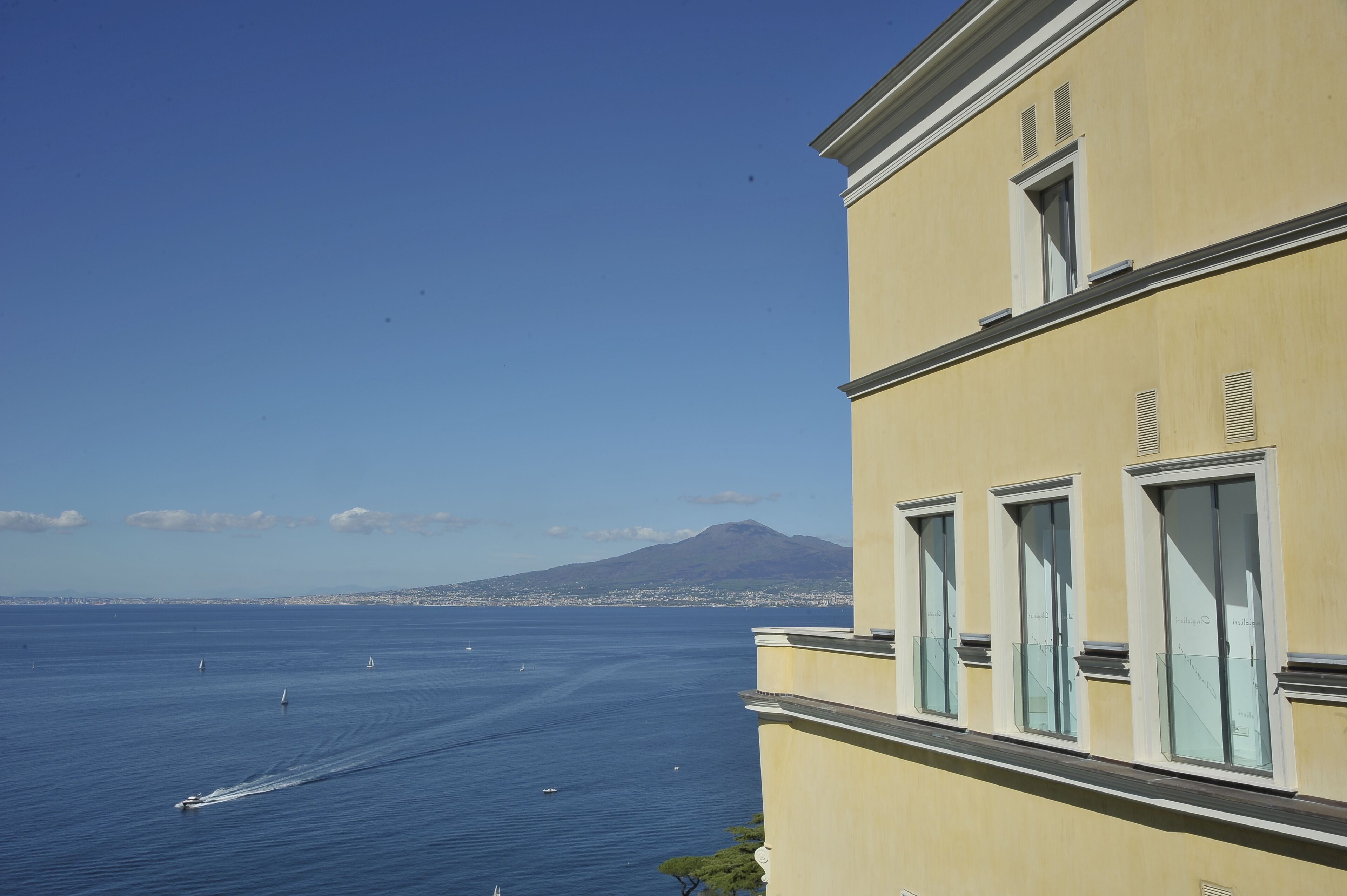 Un bâtiment situé sur la côte, doté de grandes fenêtres donnant sur la mer, sous un ciel dégagé, avec une montagne visible au loin.