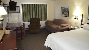 A hotel room with a bed, a desk, a chair, a sofa, a television mounted on the wall, a window with curtains, a painting on the wall, and a lamp on the nightstand. - Grand Inn Fargo (Fargo)