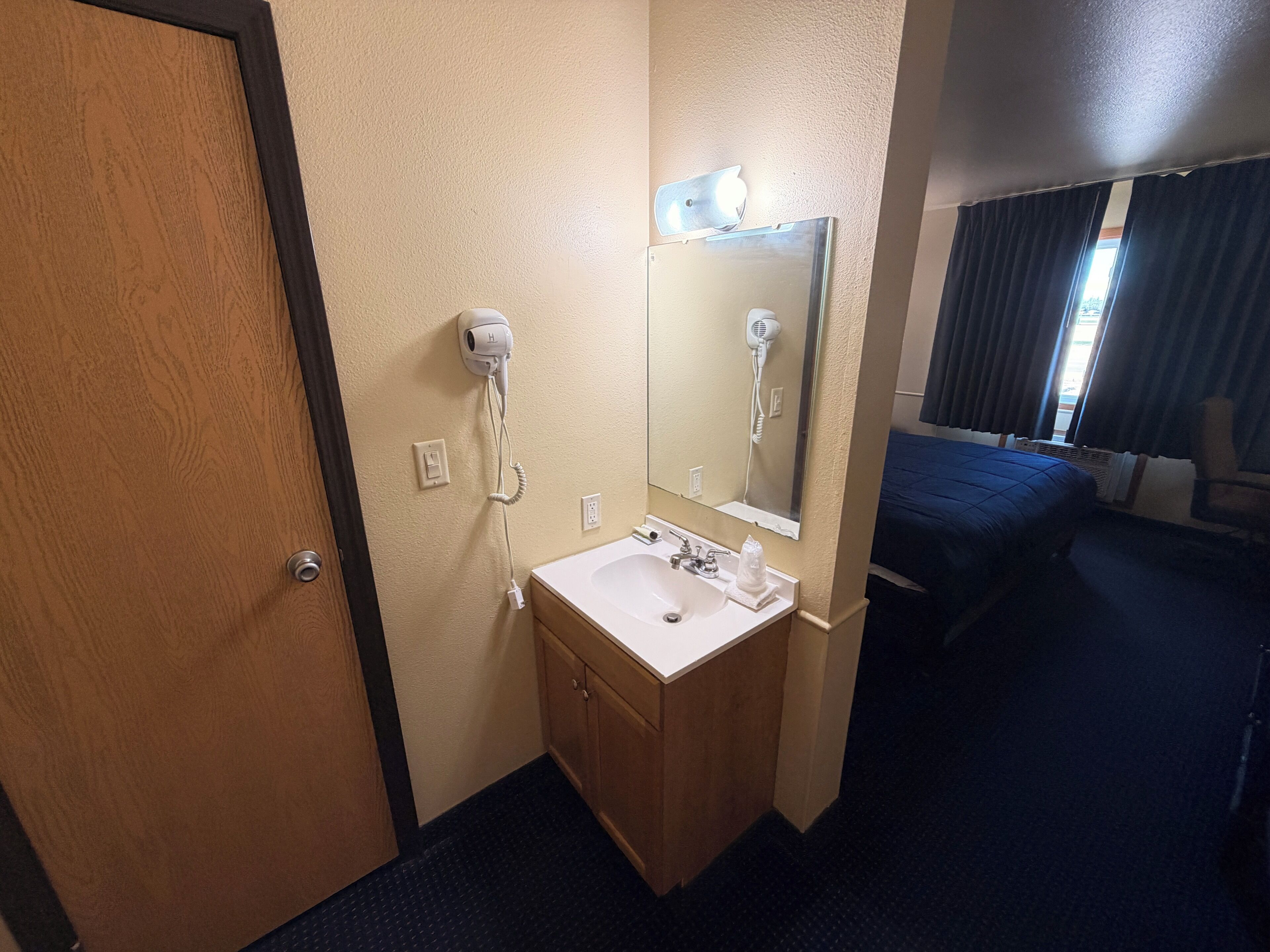 Habitación de hotel con puerta de madera, un lavabo blanco con espejo, secador de pelo, cama con cubierta azul y ventana con cortinas.
